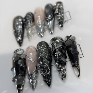 May include: A set of ten black and silver nail tips with various designs, including a cross, a star, and a wing. The tips are decorated with silver and black glitter.