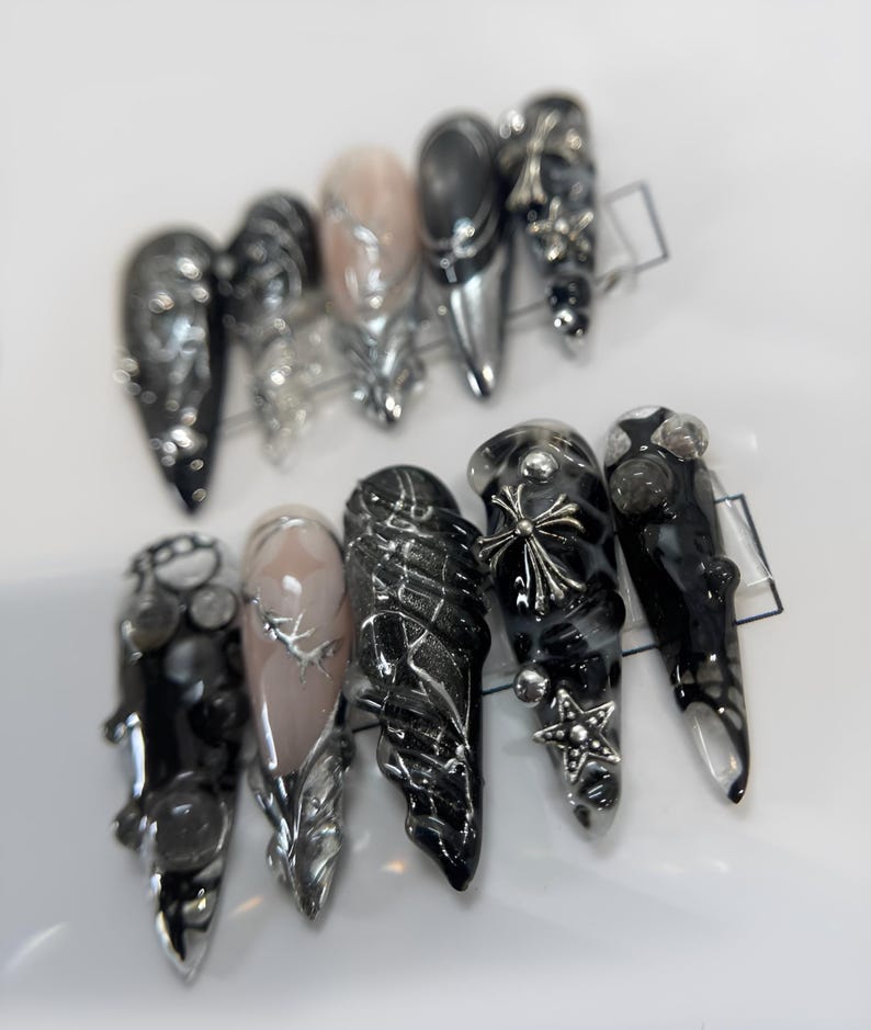 May include: A set of ten black and silver nail tips with various designs, including a cross, a star, and a wing. The tips are decorated with silver and black glitter.
