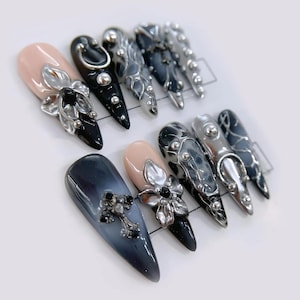 Puede incluir: Una colección de uñas artificiales con varios diseños. Algunas uñas son nude, negras o degradadas en azul-gris, adornadas con adornos florales o en forma de cruz plateados. Otras uñas presentan detalles metálicos plateados y puntas.