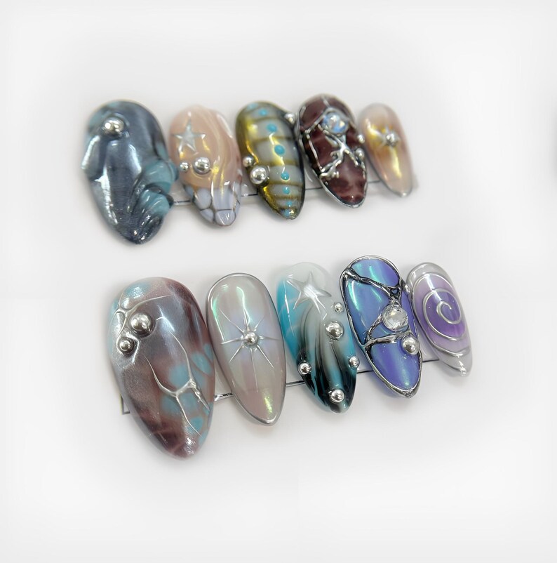 May include: A set of ten artificial nails in various designs. The nails feature a range of colors, including blue, brown, and iridescent hues. Some nails have silver embellishments and intricate patterns, while others have a smooth, polished finish.