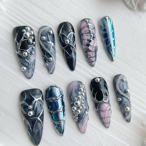 May include: A collection of ten long, almond-shaped artificial nails. Each nail features a unique design with dark, swirling colors, silver accents, and intricate patterns. Some nails have a pink and black web design, while others have silver beads.