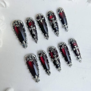 May include: A collection of ten coffin-shaped false nails. Each nail has a deep red gradient design, complemented by silver-coloured embellishments. The embellishments feature crosses, stars, and floral patterns, creating a gothic aesthetic.