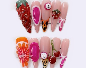 Könnte beinhalten: Ein Satz von zehn langen, mandelförmigen Kunstnägeln. Jeder Nagel hat ein anderes Design, darunter eine Erdbeere, ein rosa Oval, eine Billardkugel, eine kleine Erdbeere, eine Zitrusfrucht, ein rosa Oval, eine Billardkugel, Kirschen und eine Schleife.