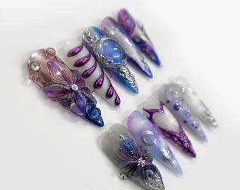 May include: A set of ten long, pointed, artificial fingernails with various designs. The designs include butterflies, swirls, moons, and hearts. The nails are painted in shades of purple, blue, and white.