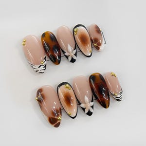 May include: Set of ten false nails with different designs. The nails are nude, brown, and black with white zebra stripes, brown tortoise shell, and white flowers. Some nails have gold accents.