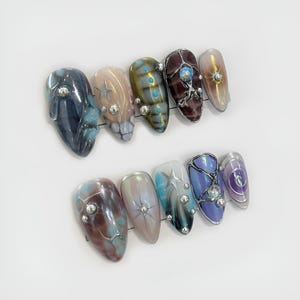 May include: A set of ten almond-shaped, press-on nails. The nails feature a variety of designs, including iridescent finishes, star accents, and jewel embellishments. Colors include blue, brown, purple, and gold.