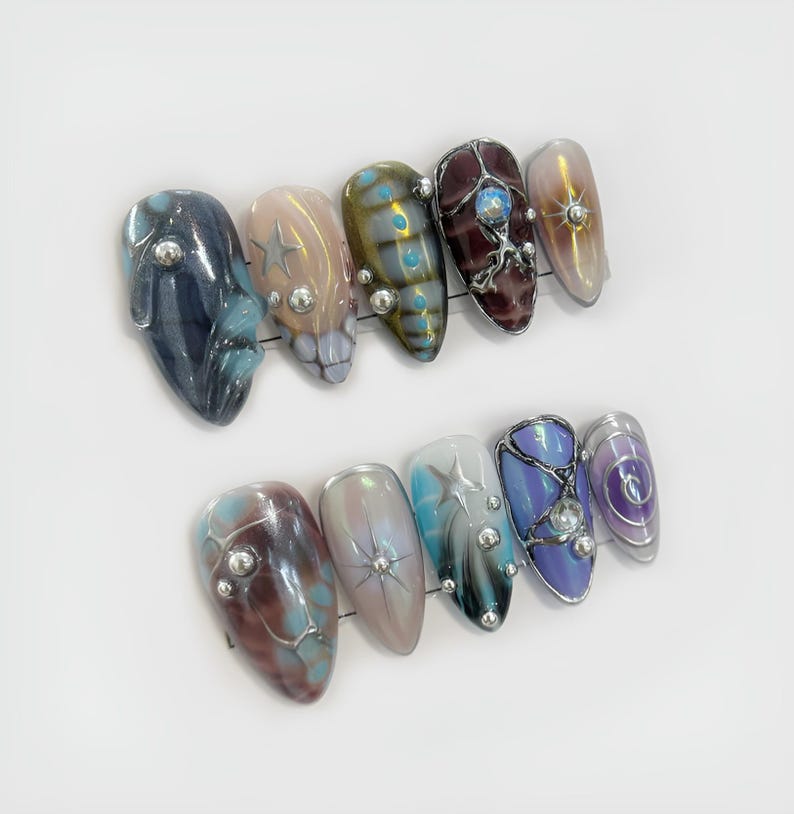 May include: A set of ten almond-shaped, press-on nails. The nails feature a variety of designs, including iridescent finishes, star accents, and jewel embellishments. Colors include blue, brown, purple, and gold.