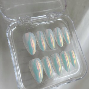 May include: A set of ten iridescent, oval-shaped false nails in a clear plastic case. The nails have a pearlescent finish that reflects shades of blue, green, and yellow. The case is open, displaying the full set of nails.