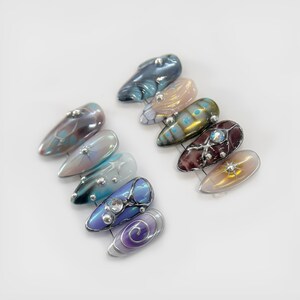 May include: A collection of ten artificial nails with various designs. The nails feature iridescent colors like blue, purple, and gold, with decorative elements such as silver studs and swirling patterns. Each nail has a glossy finish.