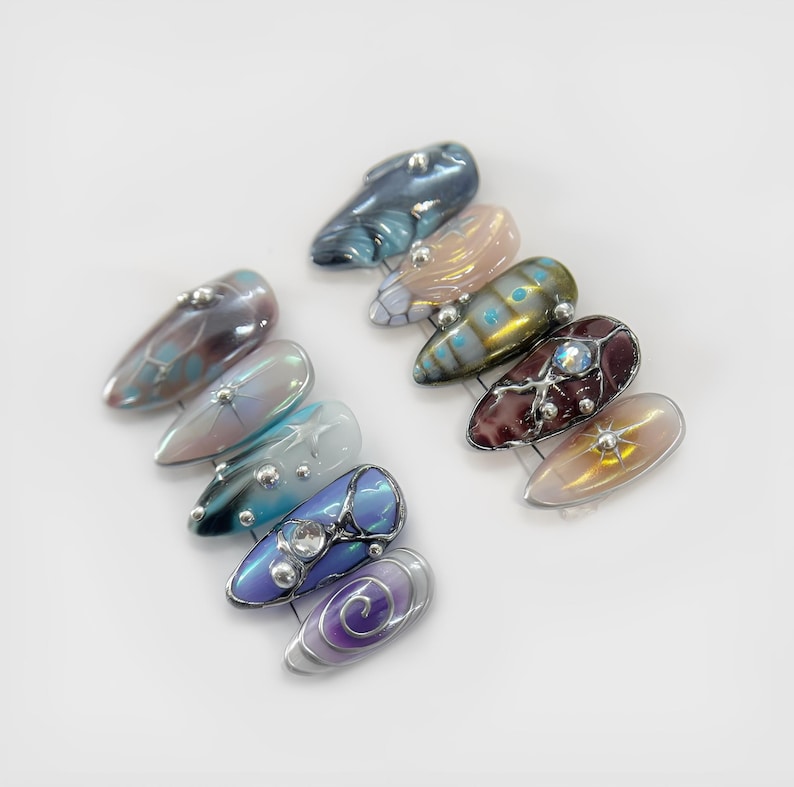 May include: A collection of ten artificial nails with various designs. The nails feature iridescent colors like blue, purple, and gold, with decorative elements such as silver studs and swirling patterns. Each nail has a glossy finish.