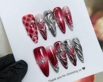 Puede incluir: Un conjunto de diez uñas artificiales largas en forma de almendra. Las uñas presentan una variedad de diseños, incluyendo un degradado rojo y blanco, acentos plateados en forma de corazón y remolinos metálicos plateados. El texto "Thank you for choosing us" es visible.