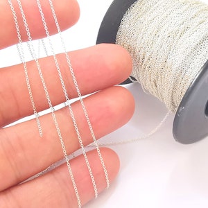 May include: A spool of silver chain, with several strands of the chain laid out on a hand. The chain is a delicate, fine gauge, and has a smooth, polished finish.
