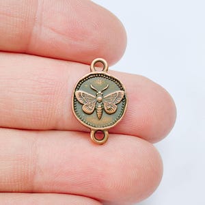 May include: A copper-colored metal charm with a moth design. The charm is round with a detailed moth in the center, surrounded by a decorative border. It has loops at the top and bottom for attaching to jewelry or other crafts.