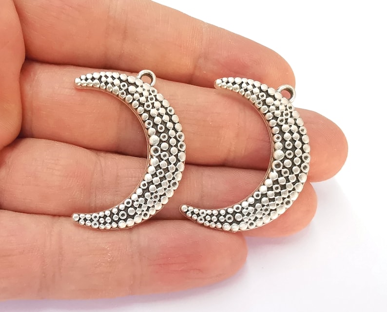 Puede incluir: Dos colgantes plateados con forma de media luna. Cada colgante tiene una superficie texturizada con peque&ntilde;os puntos en relieve. Los colgantes tienen un lazo en la parte superior para sujetarlos a la joyer&iacute;a. El fondo es blanco.