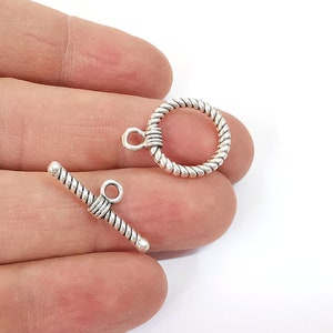 May include: Silver tone toggle clasp with a twisted rope design. The clasp has a round ring and a bar with a loop on one end.