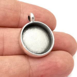 Op de afbeelding: Zilverkleurige hanger met een ronde, open achterkant voor een cameo of andere versiering. De hanger heeft een lus aan de bovenkant om aan een ketting of koord te bevestigen.