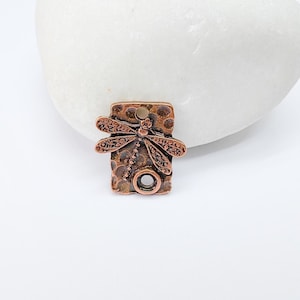 May include: Copper charm featuring a dragonfly design on a textured background. The charm has a hole for attaching to jewellery or other crafts.
