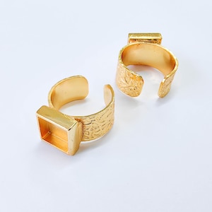May include: Two gold-colored adjustable rings. Each ring has a textured band and a rectangular setting. The rings are open-ended, allowing for size adjustment. The rings are displayed on a white surface.
