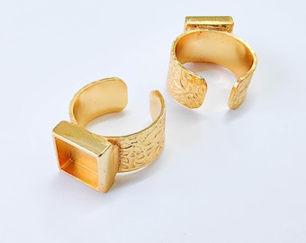 May include: Two gold-colored adjustable rings. Each ring has a textured band and a rectangular setting. The rings are open-ended, allowing for size adjustment. The rings are displayed on a white surface.
