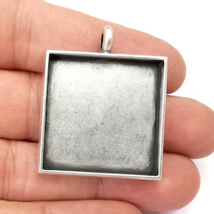Może przedstawiać: Srebrny kwadratowy wisiorek o gładkiej, lekko fakturowanej powierzchni. Wisiorek ma pętelkę u góry do przymocowania łańcuszka.