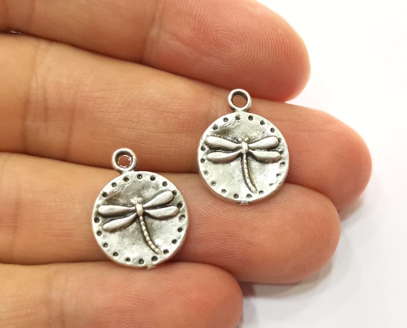 May include: Two silver-toned metal charms in the shape of a circle with a dragonfly design. The dragonfly is raised and detailed.