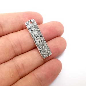 Pu&ograve; includere: Ciondolo in metallo color argento con un design floreale ornato. Il ciondolo &egrave; rettangolare e ha un foro nella parte superiore per essere attaccato a una collana o un braccialetto.