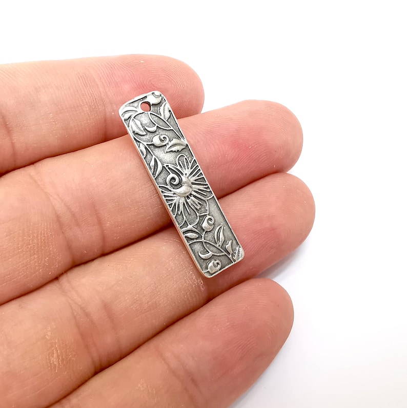 Pu&ograve; includere: Ciondolo in metallo color argento con un design floreale ornato. Il ciondolo &egrave; rettangolare e ha un foro nella parte superiore per essere attaccato a una collana o un braccialetto.