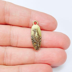 Puede incluir: Un dije de metal dorado en forma de óvalo con una imagen detallada de un pájaro posado en una rama. El dije tiene un bucle en la parte superior para sujetarlo a un collar o pulsera.