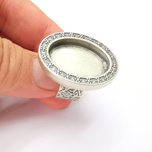 Può includere: Anello in argento con una lunetta di forma ovale, perfetto per aggiungere un ciondolo o una pietra personalizzata. La lunetta è circondata da un bordo decorativo di piccoli fiori.