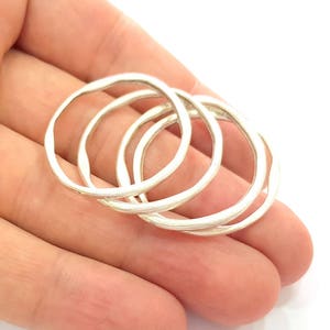 Op de afbeelding: Vier zilverkleurige ringen met een onregelmatige, organische vorm, in elkaar grijpend. De ringen hebben een getextureerd oppervlak. De ringen worden in een hand gehouden tegen een witte achtergrond.