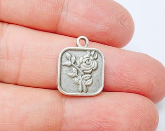 Puede incluir: Un pequeño dije cuadrado de color plateado con un diseño de rosa en relieve. El dije presenta una rosa detallada con hojas y un pequeño lazo en la parte superior para sujetarlo a un collar o pulsera. El dije se sostiene en una mano.