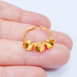 Op de afbeelding: Een goudkleurige ring met drie cirkelvormige zettingen. De ring heeft een gebogen band met kleine balaccenten aan de uiteinden. De zettingen zijn ontworpen om kleine items vast te houden. De ring wordt in de hand van een persoon gehouden.