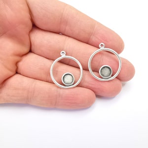 May include: Two silver-tone pendant blanks. Each features a circular frame with a small, round, recessed area for setting a stone. The pendants have a small loop at the top for attaching to a chain. Jewellery making supplies.