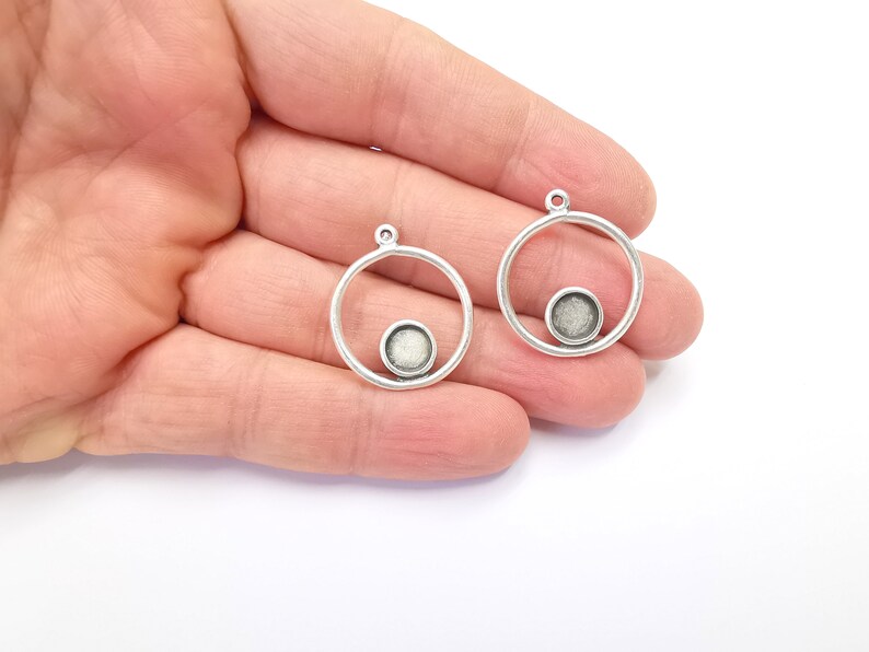 May include: Two silver-tone pendant blanks. Each features a circular frame with a small, round, recessed area for setting a stone. The pendants have a small loop at the top for attaching to a chain. Jewellery making supplies.