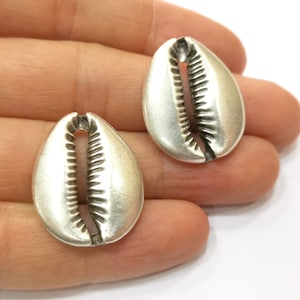 May include: Two silver cowrie shells, a type of sea shell, are shown in a hand. The shells are smooth and have a detailed, carved design.
