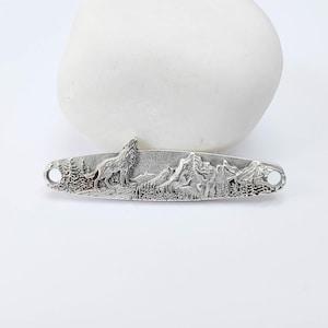 Könnte beinhalten: Ein silberfarbener Metallstab mit einer detaillierten Berg- und Waldszene, die einen heulenden Wolf zeigt. Der Stab hat an jedem Ende zwei Löcher. Das Design ist ein Relief.