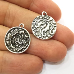 May include: Two silver-toned round pendants. One pendant features a detailed rose and floral design, while the other showcases an intricate swirling leaf pattern. Both pendants have a small loop at the top for attaching to a chain or other jewelry.