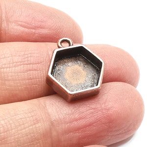 May include: A small, copper-coloured hexagonal pendant with a loop at the top. The inside of the pendant has a dark, textured surface. The pendant is held in a person's hand against a white background.