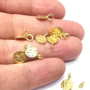 Può includere: Diversi piccoli ciondoli in metallo color oro con una texture martellata e un anello per il fissaggio ai gioielli. I ciondoli sono rotondi e hanno un piccolo perno sul fondo. I ciondoli sono sparsi su uno sfondo bianco.