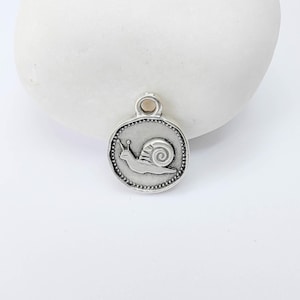 Könnte beinhalten: Ein silberfarbener Anhänger mit einem Schneckenmotiv. Der runde Anhänger hat einen Perlenrand und eine kleine Schlaufe oben. Die Schnecke ist im Profil dargestellt, mit ihrem Gehäuse und ihren Fühlern. Ideal für Schmuck.