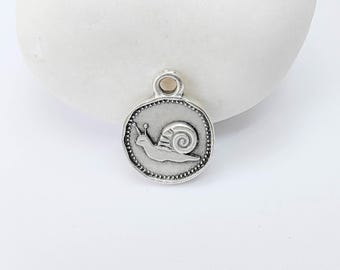 May include: A silver-colored charm with a snail design. The charm is round with a beaded border and a small loop at the top for attaching to a necklace or bracelet. The snail is depicted in profile, with its shell and antennae visible.