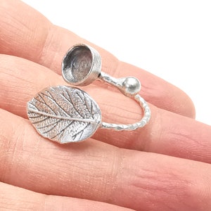 Può includere: Un anello d'argento con un design a foglia e uno spazio rotondo aperto sul lato. L'anello è su un dito.