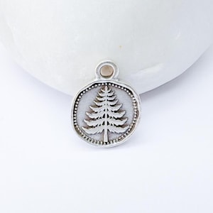 May include: A silver-colored pendant with a detailed tree design. The pendant is round with a dotted border and a small loop at the top for a chain. The tree is centered on a textured background.