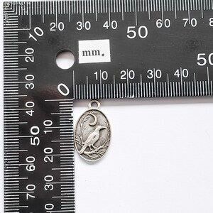 May include: A silver charm featuring a detailed design of a crow perched on a branch with a crescent moon in the background. The charm is approximately 2 cm in diameter.