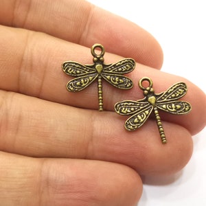 Può includere: Due ciondoli a forma di libellula dorati antichi con dettagli intricati. I ciondoli hanno un anello nella parte superiore per essere attaccati a gioielli o altri oggetti artigianali.