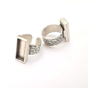 May include: Two silver rings with rectangular settings for adding a stone or other embellishment. The rings have a textured band.