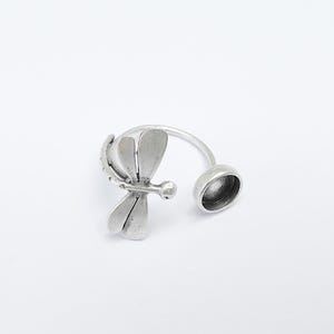 May include: Silver ring with a dragonfly design and a round bezel setting. The ring is open and adjustable.