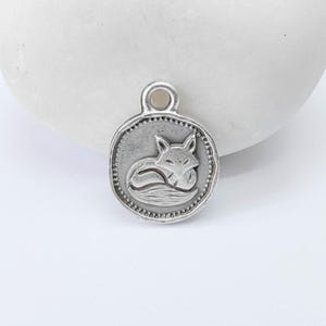 Op de afbeelding: Zilverkleurige hanger met een vosontwerp. De ronde hanger heeft een gedetailleerde rand en een lus aan de bovenkant om op te hangen. De vos is opgerold afgebeeld, wat een speels tintje aan de sieraden geeft.