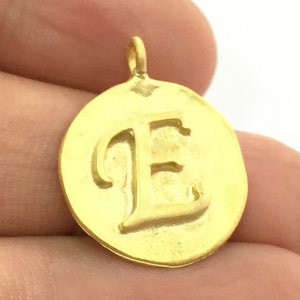 May include: Gold-toned pendant charm featuring the letter E in a cursive font. The charm is round and has a smooth, polished finish.