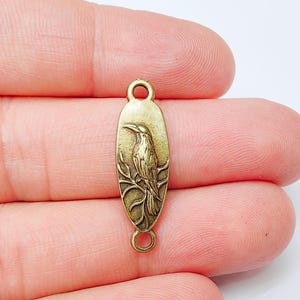 May include: A bronze-coloured metal charm with a bird design. The charm is oval-shaped with a detailed relief of a bird perched on a branch. It has a loop at the top and bottom for attaching to jewellery or other crafts.